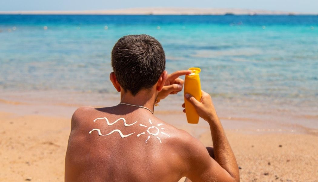 Man sitting on a sunny beach, holding a yellow sunscreen bottle and applying sunscreen to his back, where a sun and wave design is drawn in lotion—highlighting the importance of sun protection as a key factor in reducing skin cancer risk.