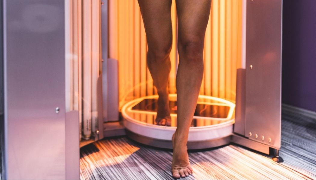 A person stepping out of a vertical indoor tanning booth, with orange UV lights illuminating the interior, illustrating the risks associated with indoor tanning, such as increased chances of skin cancer, premature aging, and UV skin damage.